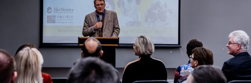 A conference room full of people, in the middle a man with a microphone is standing: the director of the museum welcoming guests. In the background there is a screen with the title of the exhibition &quot;In the footsteps of Jewish women in Europe&quot;
