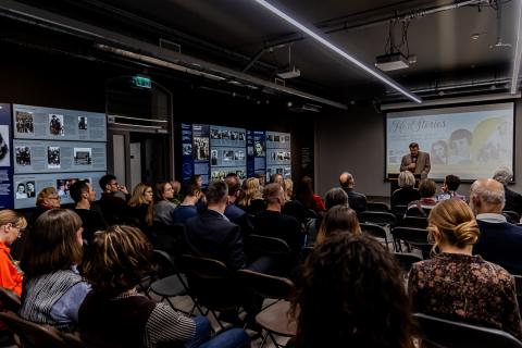 A conference room full of people, in the middle a man with a microphone is standing: the director of the museum welcoming guests. In the background there is a screen with the title of the exhibition "In the footsteps of Jewish women in Europe"