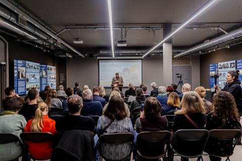 A conference room full of people, in the middle a man with a microphone is standing: the director of the museum welcoming guests. In the background there is a screen with the title of the exhibition "In the footsteps of Jewish women in Europe"