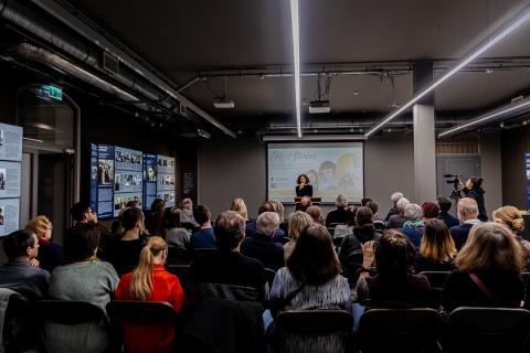 A conference room full of people. A woman - curator of the exhibition - is standing in front of them and speaking.