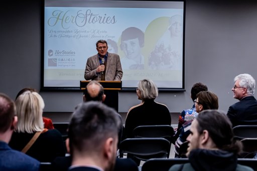 A conference room full of people, in the middle a man with a microphone is standing: the director of the museum welcoming guests. In the background there is a screen with the title of the exhibition "In the footsteps of Jewish women in Europe"